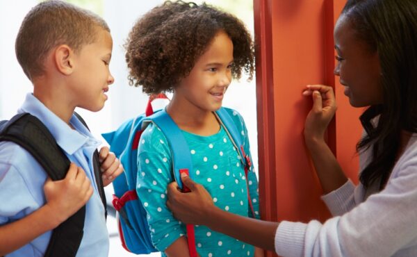 An older black woman sending off two children at the door. The kids wear backpacks and smile at the woman.