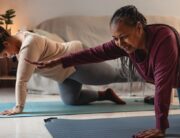 A Black mother and daughter are smiling as they hold a yoga pose together in a dimly lit living room.