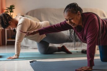 A Black mother and daughter are smiling as they hold a yoga pose together in a dimly lit living room.