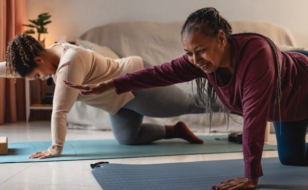 A Black mother and daughter are smiling as they hold a yoga pose together in a dimly lit living room.