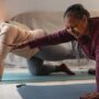 A Black mother and daughter are smiling as they hold a yoga pose together in a dimly lit living room.