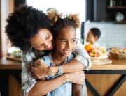A Black mother and her daughter are sitting in the kitchen, with the mom hugging her daughter from behind.