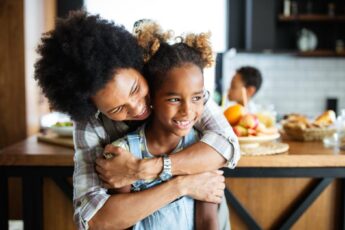 A Black mother and her daughter are sitting in the kitchen, with the mom hugging her daughter from behind.