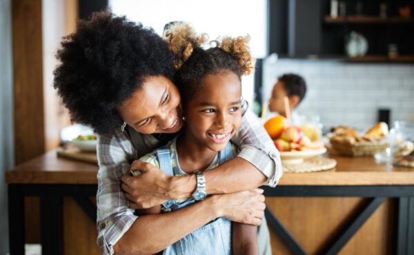 A Black mother and her daughter are sitting in the kitchen, with the mom hugging her daughter from behind.