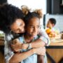A Black mother and her daughter are sitting in the kitchen, with the mom hugging her daughter from behind.