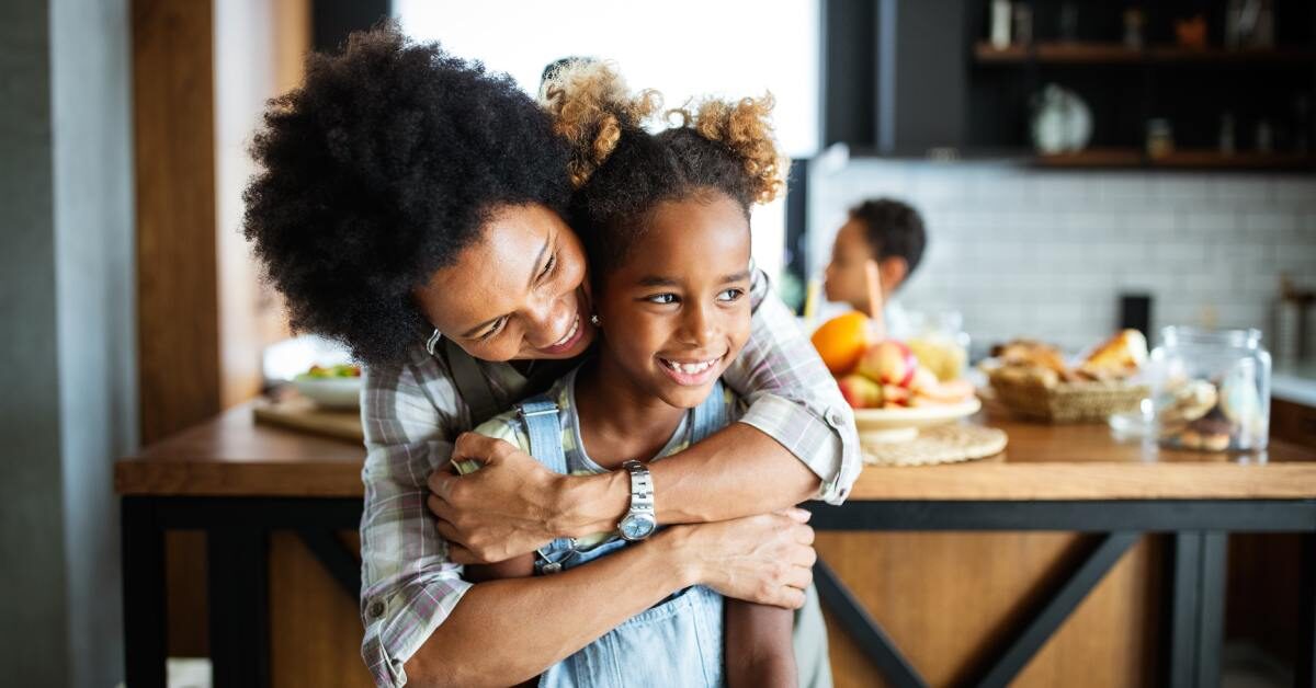 A Black mother and her daughter are sitting in the kitchen, with the mom hugging her daughter from behind.