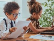 Adorable boy and girl are pretending to be professionals. The girl is typing on a laptop while the boy reads from a paper.