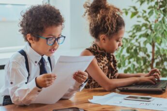 Adorable boy and girl are pretending to be professionals. The girl is typing on a laptop while the boy reads from a paper.