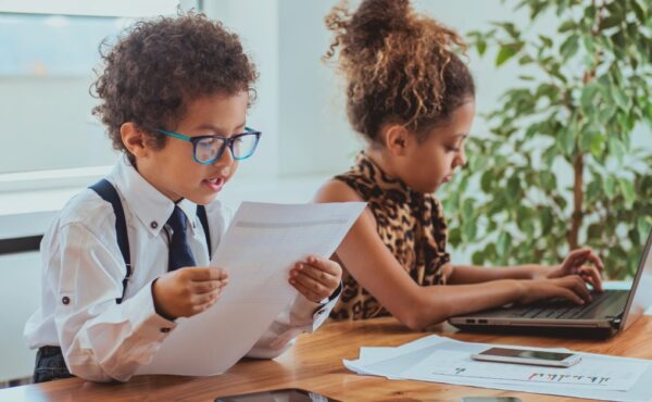 Adorable boy and girl are pretending to be professionals. The girl is typing on a laptop while the boy reads from a paper.