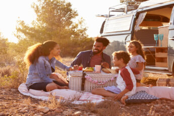 A man and woman sit smiling around a picnic basket with a young boy and girl. They are next to a blue van.