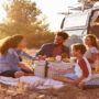 A man and woman sit smiling around a picnic basket with a young boy and girl. They are next to a blue van.