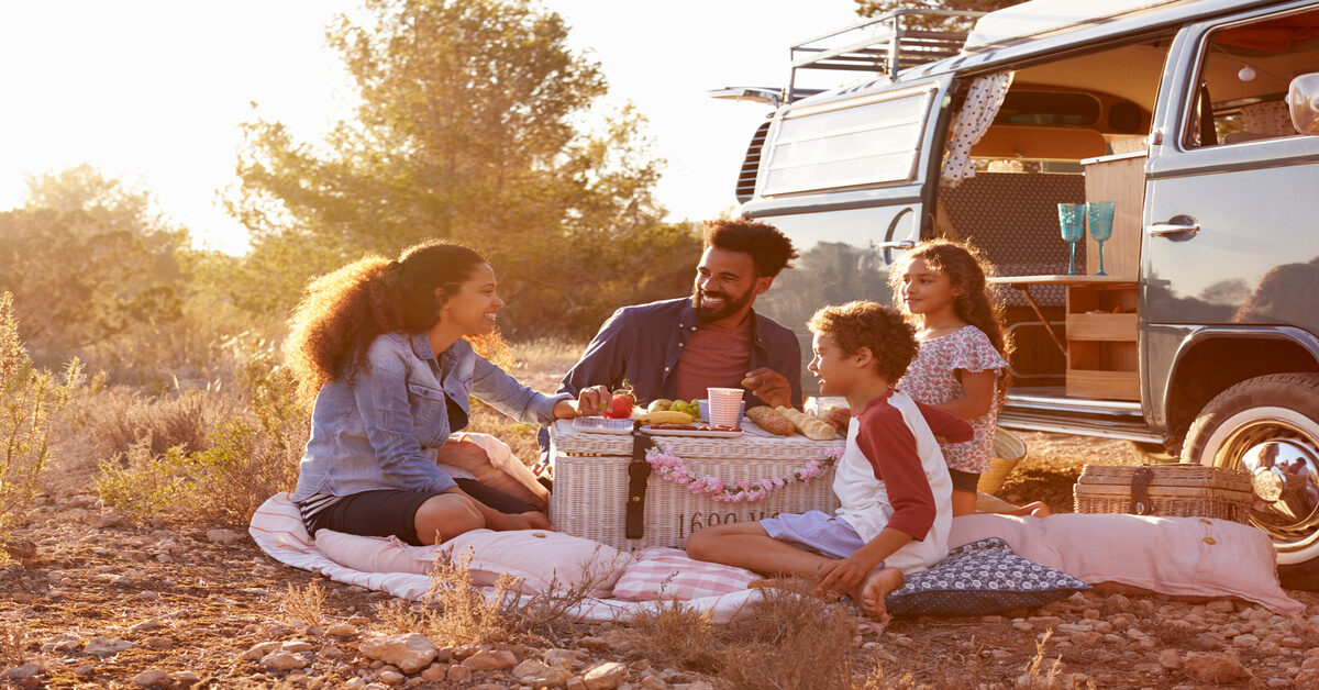 A man and woman sit smiling around a picnic basket with a young boy and girl. They are next to a blue van.