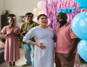 A pregnant woman and a man smile at blue balloons as a crowd claps behind them at a gender reveal party.