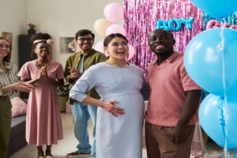 A pregnant woman and a man smile at blue balloons as a crowd claps behind them at a gender reveal party.