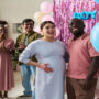 A pregnant woman and a man smile at blue balloons as a crowd claps behind them at a gender reveal party.