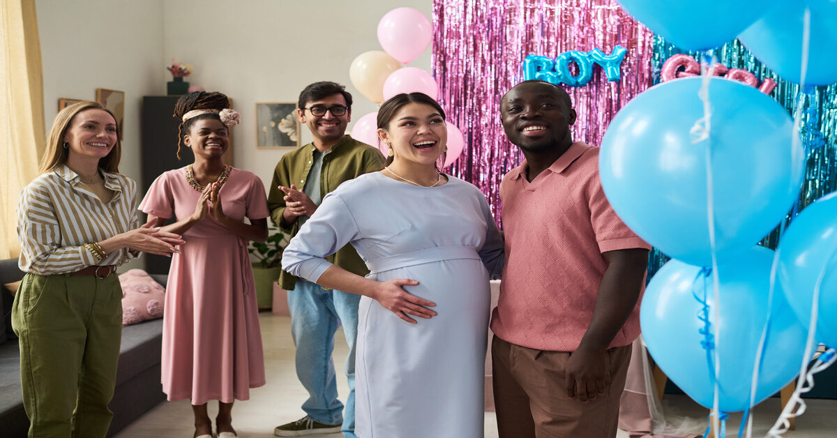 A pregnant woman and a man smile at blue balloons as a crowd claps behind them at a gender reveal party.