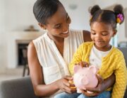 A young Black mother smiles down at her daughter, who is sitting on her lap and looking at the piggy bank in her mom’s hand.