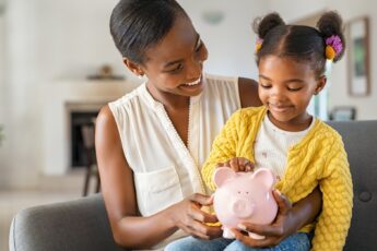 A young Black mother smiles down at her daughter, who is sitting on her lap and looking at the piggy bank in her mom’s hand.