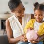 A young Black mother smiles down at her daughter, who is sitting on her lap and looking at the piggy bank in her mom’s hand.