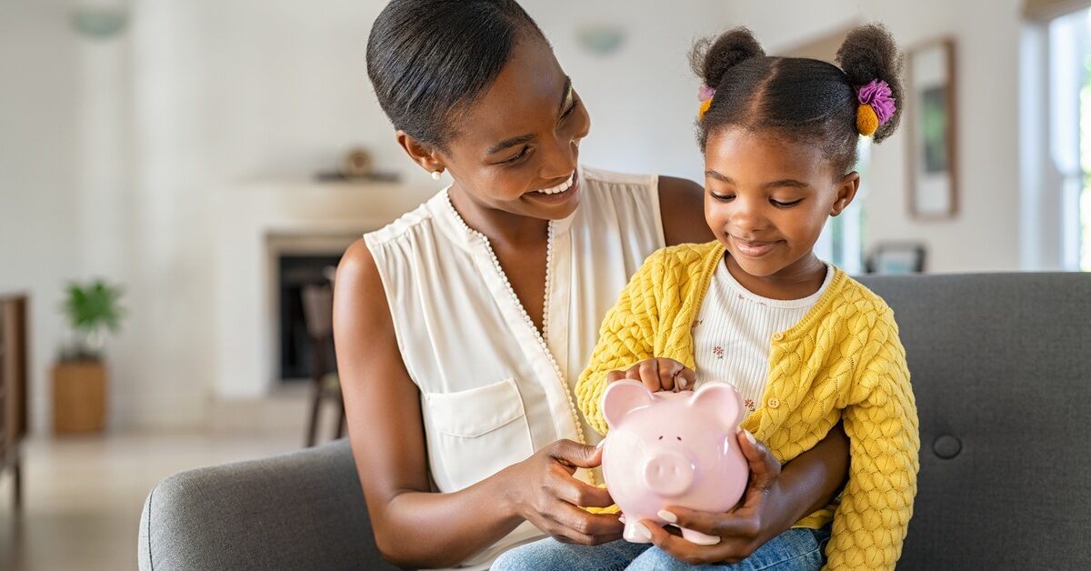 A young Black mother smiles down at her daughter, who is sitting on her lap and looking at the piggy bank in her mom’s hand.