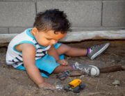 A little boy plays in a sandbox with his toy cars. The area features logs as barriers and sits next to a concrete wall.