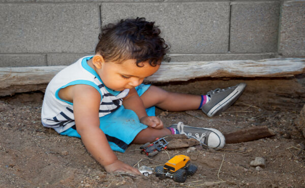 A little boy plays in a sandbox with his toy cars. The area features logs as barriers and sits next to a concrete wall.