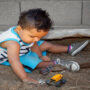A little boy plays in a sandbox with his toy cars. The area features logs as barriers and sits next to a concrete wall.