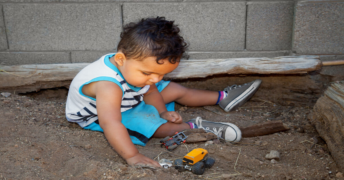 A little boy plays in a sandbox with his toy cars. The area features logs as barriers and sits next to a concrete wall.