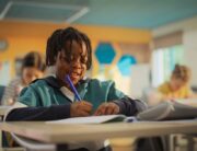 A young Black boy sits at his desk in the classroom as he writes with a blue utensil. He's writing and smiling.