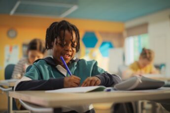 A young Black boy sits at his desk in the classroom as he writes with a blue utensil. He's writing and smiling.