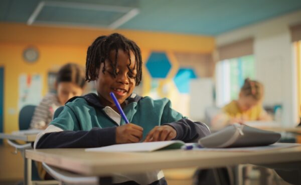 A young Black boy sits at his desk in the classroom as he writes with a blue utensil. He's writing and smiling.
