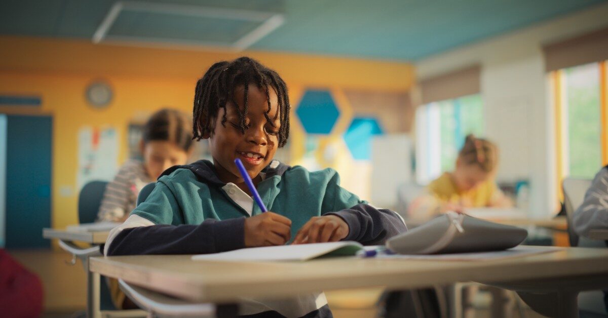 A young Black boy sits at his desk in the classroom as he writes with a blue utensil. He's writing and smiling.