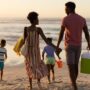 A young, Black family and their two kids are running happily towards the shoreline of the beach with toys and floaties.