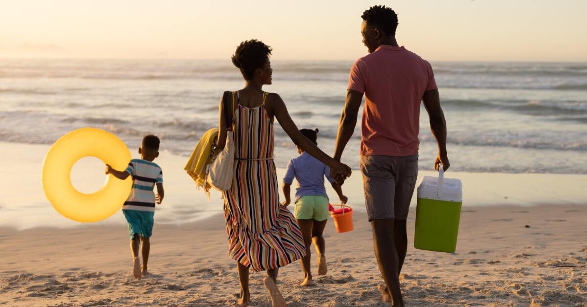 A young, Black family and their two kids are running happily towards the shoreline of the beach with toys and floaties.