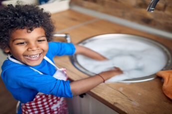 A Black toddler stands happily at the kitchen sink with an apron on, ready to help with dishes and chores.