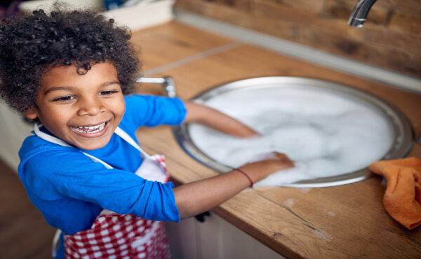 A Black toddler stands happily at the kitchen sink with an apron on, ready to help with dishes and chores.