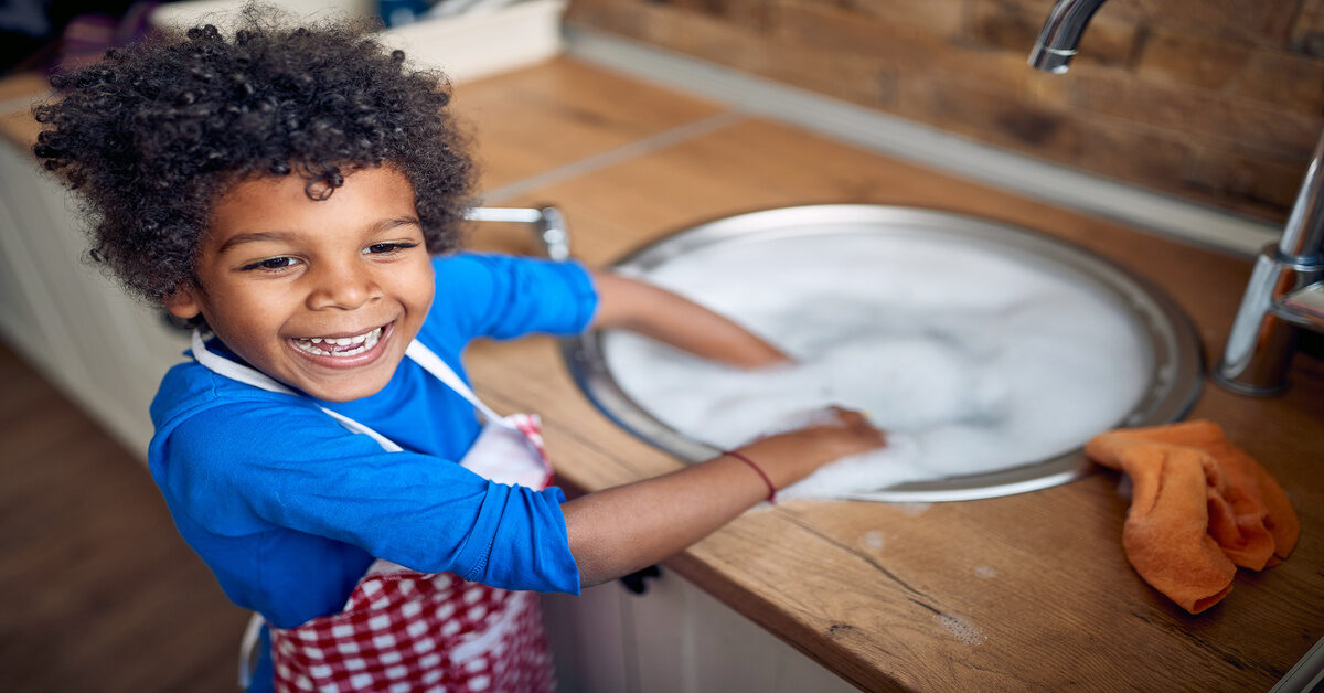 A Black toddler stands happily at the kitchen sink with an apron on, ready to help with dishes and chores.