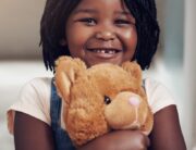 A little Black girl with braids and a missing front tooth grins as she hugs a latte-colored teddy bear to her chest.