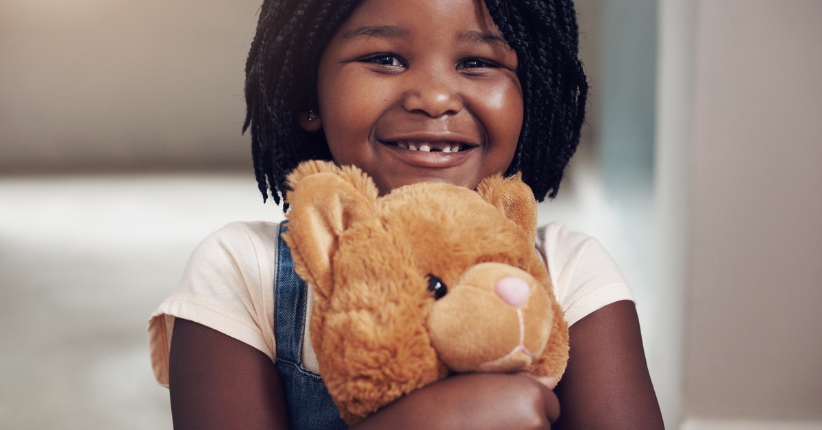 A little Black girl with braids and a missing front tooth grins as she hugs a latte-colored teddy bear to her chest.