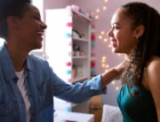 A Black mother and her daughter are smiling and laughing together on prom night in the daughter's room.